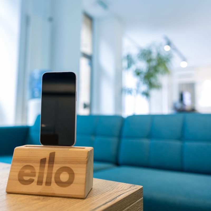 Smartphone reconditionné en exposition au ello Shop à Neuchâtel, présenté sur un support en bois avec le logo ello. Décor moderne avec un canapé bleu en arrière-plan, soulignant l'engagement d'ello pour la durabilité et l'élégance.